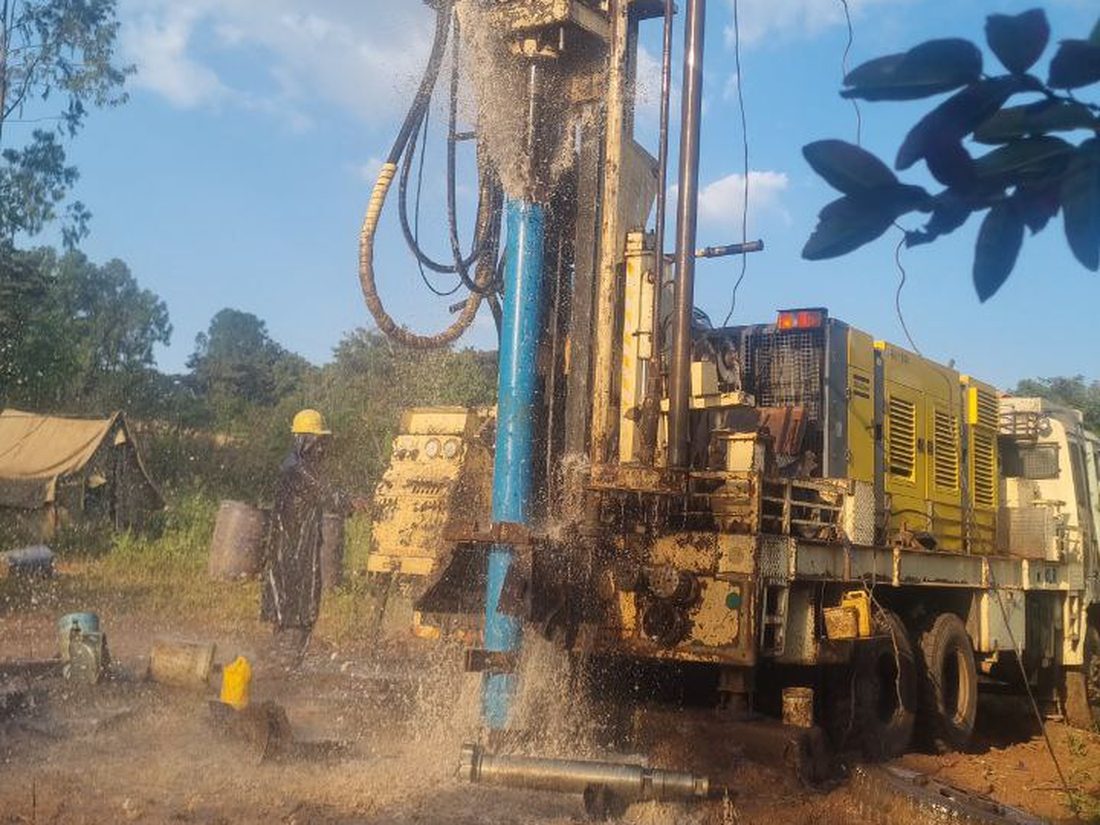 A yellow rig truck and hard-hat crew with water and cuttings spraying mid-drill